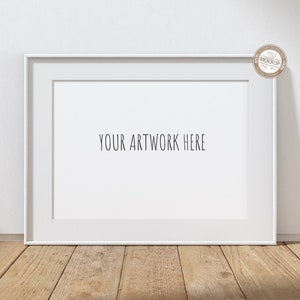May include: White rectangular picture frame with a white mat, resting on a wooden surface. The text "YOUR ARTWORK HERE" is centered within the frame. A small, circular sticker with text is in the upper right corner.