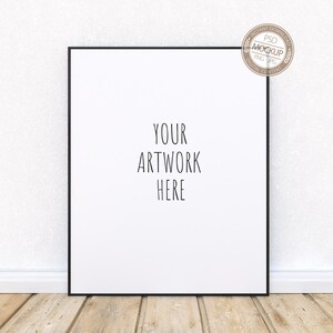 May include: A black framed poster mockup with the text "YOUR ARTWORK HERE" on a white background. The poster is leaning against a white wall with a white baseboard and wood floor.