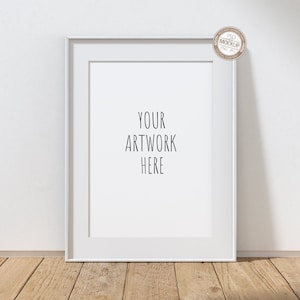 Puede incluir: Maqueta de marco de imagen blanco sobre un suelo de madera con una pared blanca de fondo. El marco está vacío y tiene el texto "YOUR ARTWORK HERE" escrito en el centro.