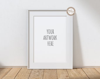 May include: White picture frame mockup on a wooden floor with a white wall in the background. The frame is empty and has the text "YOUR ARTWORK HERE" written in the center.