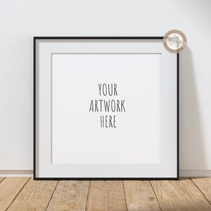 May include: Square black picture frame with a white mat, displaying the text "YOUR ARTWORK HERE." The frame is set against a white wall and rests on a wooden surface. A small circular logo is in the upper right corner.