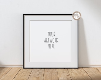 Könnte beinhalten: Quadratischer schwarzer Bilderrahmen mit weißem Passepartout, der den Text "YOUR ARTWORK HERE" anzeigt. Der Rahmen steht vor einer weißen Wand und ruht auf einer Holzoberfläche. Ein kleines kreisförmiges Logo befindet sich in der oberen rechten Ecke.