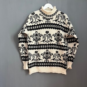 Könnte beinhalten: Ein cremefarbener Strickpullover mit einem schwarz-weißen geometrischen Muster. Das Muster zeigt stilisierte Bäume, Vögel und geometrische Formen.