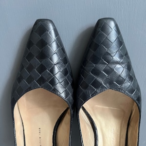 Puede incluir: Un par de tacones negros de cuero con tira trasera y un patrón tejido. Los tacones tienen una punta puntiaguda y un tacón bajo. Los zapatos están etiquetados "Peter Kaiser since 1838."