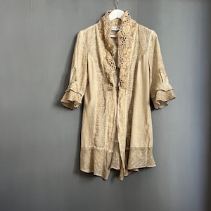 Könnte beinhalten: Eine beige Leinenjacke mit Spitzenkragen und Rüschenärmeln. Die Jacke hat einen Knopfverschluss und einen etwas längeren Rücken.