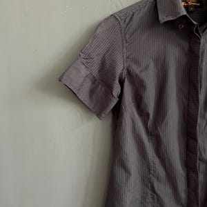 May include: A charcoal grey short-sleeved shirt with vertical pinstripes, hanging on a white hanger. The shirt features a classic collar and a tailored fit. The shirt is a dark grey colour.