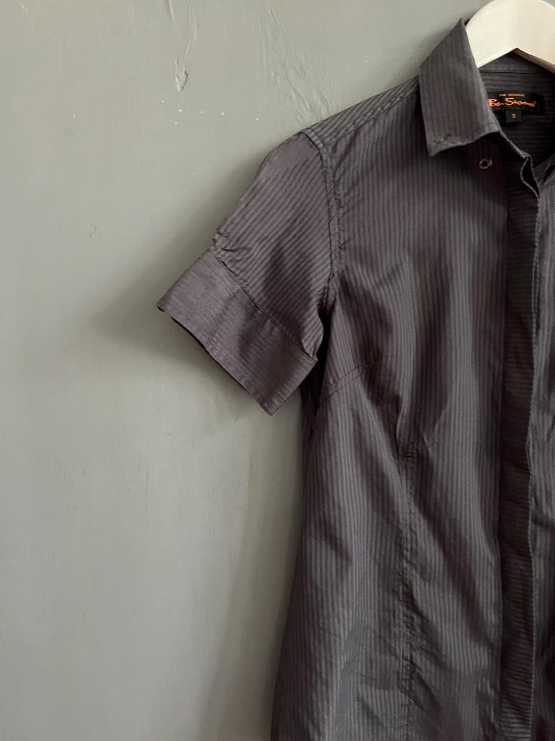 May include: A charcoal grey short-sleeved shirt with vertical pinstripes, hanging on a white hanger. The shirt features a classic collar and a tailored fit. The shirt is a dark grey colour.
