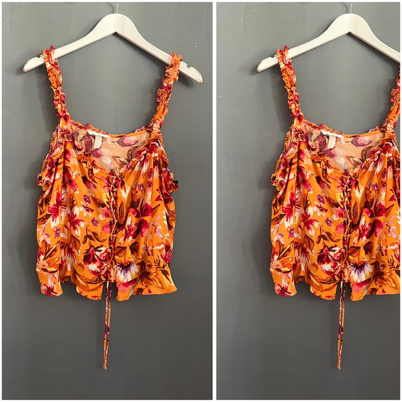 May include: An orange floral print top with ruffled straps and a lace-up front. The top is hanging on a white hanger against a gray background. The floral pattern includes red, pink, and white flowers.
