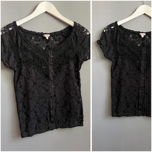 May include: Black lace top with short sleeves and a button-up front. The top is displayed on a white hanger against a grey background. The lace pattern is detailed and covers the entire garment.