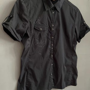 May include: A black short-sleeved button-down shirt with two chest pockets and roll-up sleeves. The shirt has a collared neckline and a series of small silver buttons down the front. A small floral design is visible on the left side of the shirt.