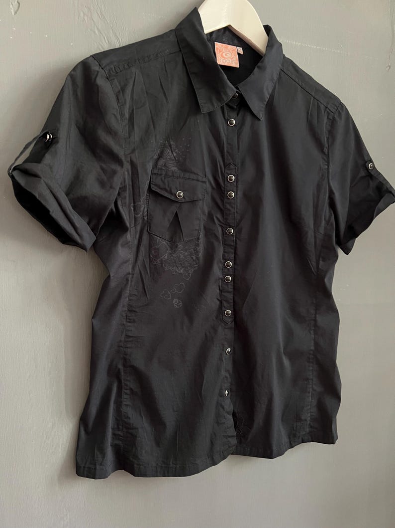 May include: A black short-sleeved button-down shirt with two chest pockets and roll-up sleeves. The shirt has a collared neckline and a series of small silver buttons down the front. A small floral design is visible on the left side of the shirt.