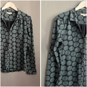 May include: A long-sleeved shirt with a dark base and a pattern of teal circles. The shirt features a collar, a chest pocket, and small, round buttons. It is displayed on a white hanger. The shirt is made of a soft material.