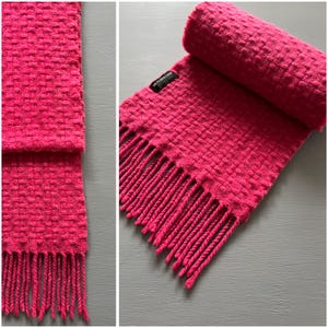 May include: A pink knitted scarf with a fringe on both ends. The scarf is rolled up and has a black tag that says "Fletcher Moss" on it.