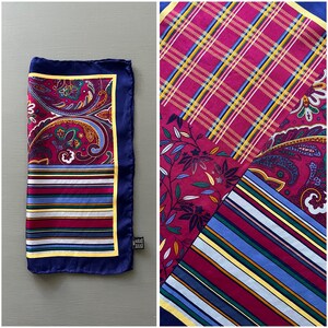 May include: A square scarf featuring a patchwork design with sections of magenta paisley, multi-colored horizontal stripes, and a plaid pattern. The scarf is bordered by a solid navy blue edge and a thin yellow frame, with a small black tag attached.