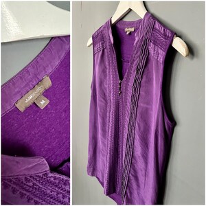 May include: A sleeveless purple blouse with a V-neck and decorative stitching down the front. The blouse has a small collar and is hanging on a white hanger. The tag reads "JJBenson" and "M".