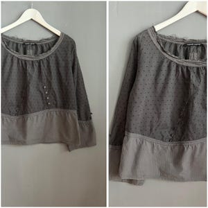 Könnte beinhalten: Graue Langarmbluse mit gepunktetem Oberteil und gestreiftem Unterteil. Die Bluse hat einen runden Ausschnitt und Knopfdetails vorne. Das Kleidungsstück hängt an einem weißen Kleiderbügel.