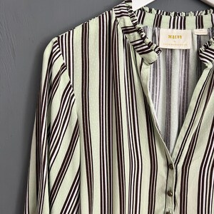 May include: A green and brown striped shirt with a ruffled collar and button closure. The shirt has a small label that reads "Maeve by Anthropologie".
