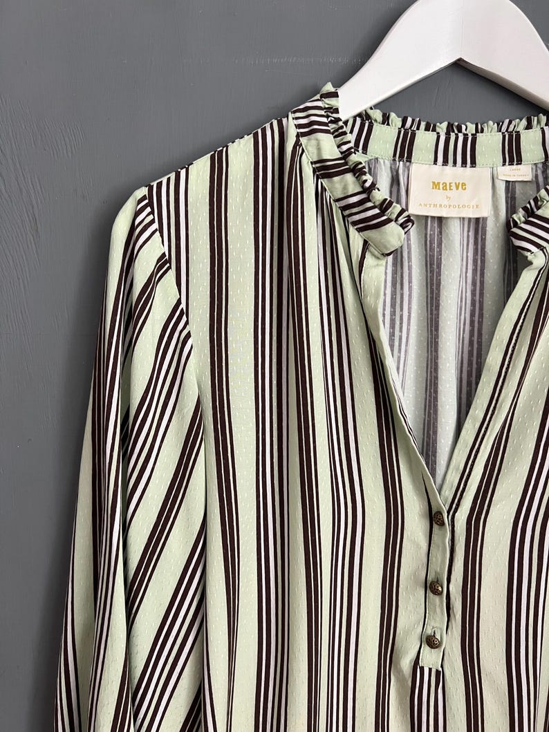 May include: A green and brown striped shirt with a ruffled collar and button closure. The shirt has a small label that reads "Maeve by Anthropologie".