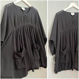 Puede incluir: Una túnica negra de algodón, holgada, con cuello redondo, cierre de botón y tres bolsillos grandes. La túnica tiene una cintura fruncida y un dobladillo ligeramente acampanado.