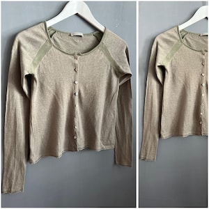May include: A long-sleeved, button-down cardigan in a neutral beige color. The cardigan features olive green accents on the shoulders and neckline. The garment is displayed on a white hanger against a gray background.