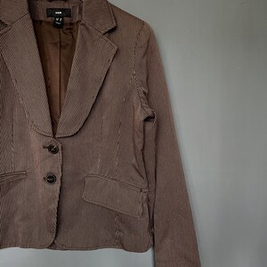 K&ouml;nnte beinhalten: Ein braunes und wei&szlig; gestreiftes Blazer mit einem Schalkragen und drei Kn&ouml;pfen. Der Blazer hat eine Tasche auf der linken Seite und ist aus einem leichten Stoff gefertigt.