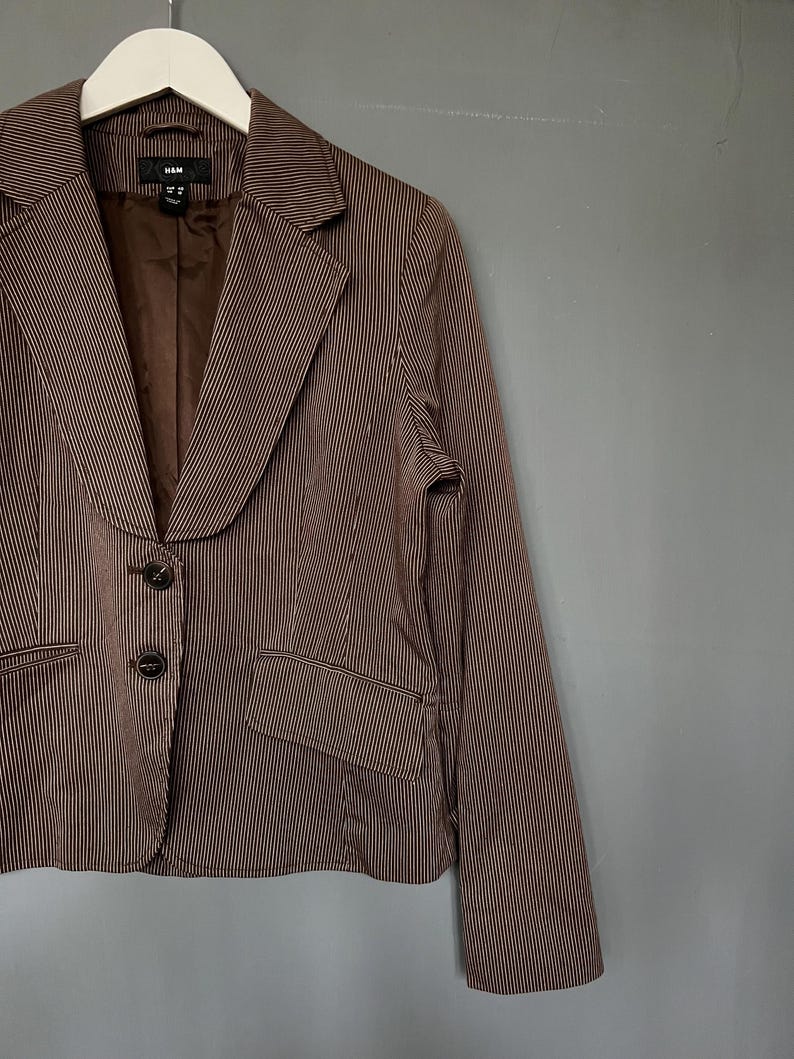K&ouml;nnte beinhalten: Ein braunes und wei&szlig; gestreiftes Blazer mit einem Schalkragen und drei Kn&ouml;pfen. Der Blazer hat eine Tasche auf der linken Seite und ist aus einem leichten Stoff gefertigt.