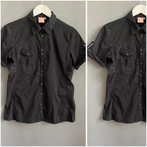 May include: Black short-sleeve button-down shirt with two chest pockets and a collared neckline. The shirt has a subtle embroidered design on the pockets and front. The sleeves can be rolled up and secured with a button. The shirt is hanging on a white hanger.
