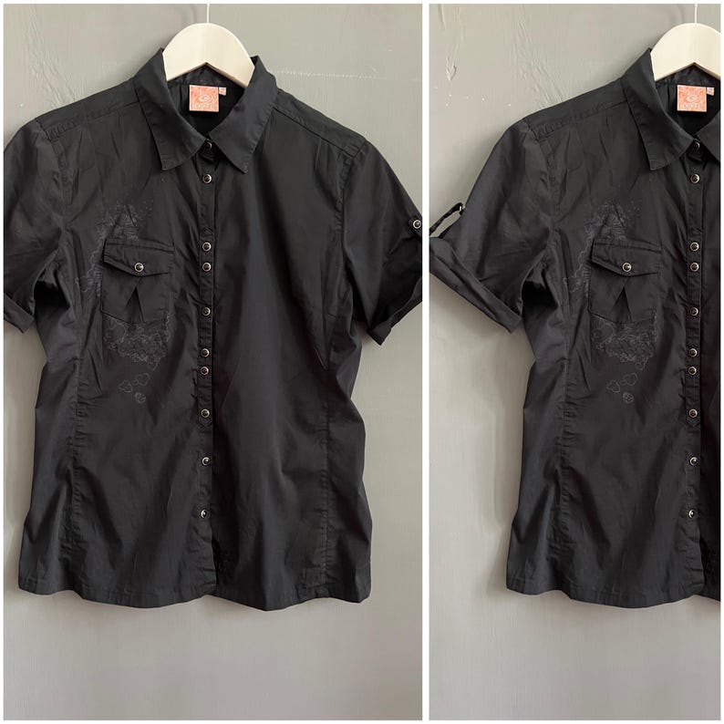 May include: Black short-sleeve button-down shirt with two chest pockets and a collared neckline. The shirt has a subtle embroidered design on the pockets and front. The sleeves can be rolled up and secured with a button. The shirt is hanging on a white hanger.
