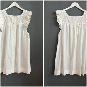 May include: White sleeveless top with a square neckline and ruffled cap sleeves. The top has small, evenly spaced white dots and a button closure at the neckline. The garment is hanging on a white hanger.