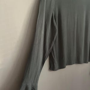 May include: A long-sleeved, sage green blouse with a high neckline and flared sleeves. The fabric has a subtle sheen, and the sleeves flare out at the wrist. The blouse is a solid color and appears to be made of a lightweight material.
