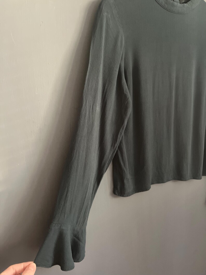 May include: A long-sleeved, sage green blouse with a high neckline and flared sleeves. The fabric has a subtle sheen, and the sleeves flare out at the wrist. The blouse is a solid color and appears to be made of a lightweight material.