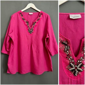 May include: A pink linen top with a beaded neckline. The neckline features a floral design with black beads and sequins. The top has a V-neck and three-quarter sleeves. The label "Candas" is visible on the top.