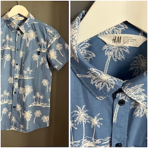 Puede incluir: Una camisa azul de manga corta con botones y estampado de palmeras blancas. La camisa tiene un bolsillo en el lado izquierdo y una etiqueta que dice "H&M" en el lado derecho.