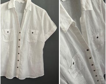 May include: White linen shirt with short sleeves, two front pockets, and black buttons. The shirt has a collared neckline and is hanging on a white hanger. The brand name "Marks & Spencer" is visible on the label.