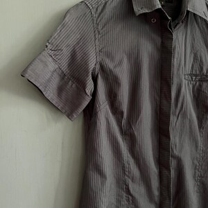 May include: A grey, short-sleeved shirt with vertical stripes. The shirt features a collar, a chest pocket, and a tailored fit. The fabric looks to be a lightweight material, suitable for warmer weather.