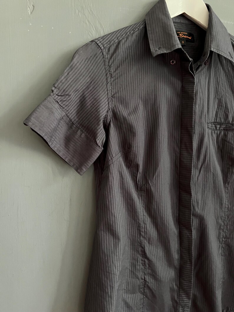 May include: A grey, short-sleeved shirt with vertical stripes. The shirt features a collar, a chest pocket, and a tailored fit. The fabric looks to be a lightweight material, suitable for warmer weather.