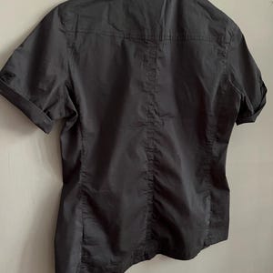May include: A black short-sleeved shirt with a collared neckline. The back of the shirt is visible, showing a vertical seam down the center and horizontal seams across the shoulders. The sleeves are cuffed.