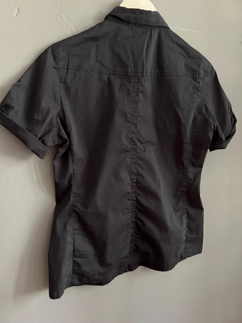 May include: A black short-sleeved shirt with a collared neckline. The back of the shirt is visible, showing a vertical seam down the center and horizontal seams across the shoulders. The sleeves are cuffed.