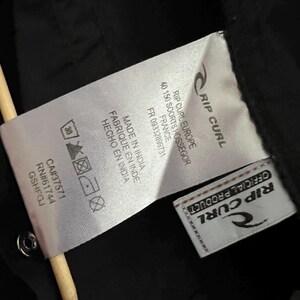 May include: Close-up of a black garment tag with the Rip Curl logo and text. The tag includes washing instructions, a CA number, and the text "MADE IN INDIA". A wooden stick is in the frame.