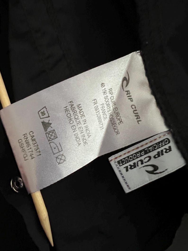 May include: Close-up of a black garment tag with the Rip Curl logo and text. The tag includes washing instructions, a CA number, and the text "MADE IN INDIA". A wooden stick is in the frame.