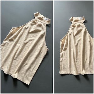 May include: A beige halter top with a textured, vertical striped pattern. The top features a high neckline and a sleeveless design. The fabric appears lightweight, suitable for warmer weather.
