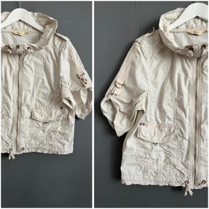 May include: A cream-colored short-sleeved jacket with a zipper closure, a hood, and two pockets. The jacket has decorative straps on the sleeves and a drawstring at the bottom.