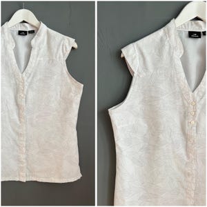 Op de afbeelding: Witte, mouwloze blouse met een V-hals en een knoopsluiting aan de voorkant. De blouse heeft een subtiel bladmotief.
