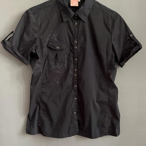 May include: Black short-sleeve button-down shirt with a collared neckline and a small pocket on the left chest. The shirt has a subtle floral design on the front and roll-up sleeves. The shirt is hanging on a white hanger.