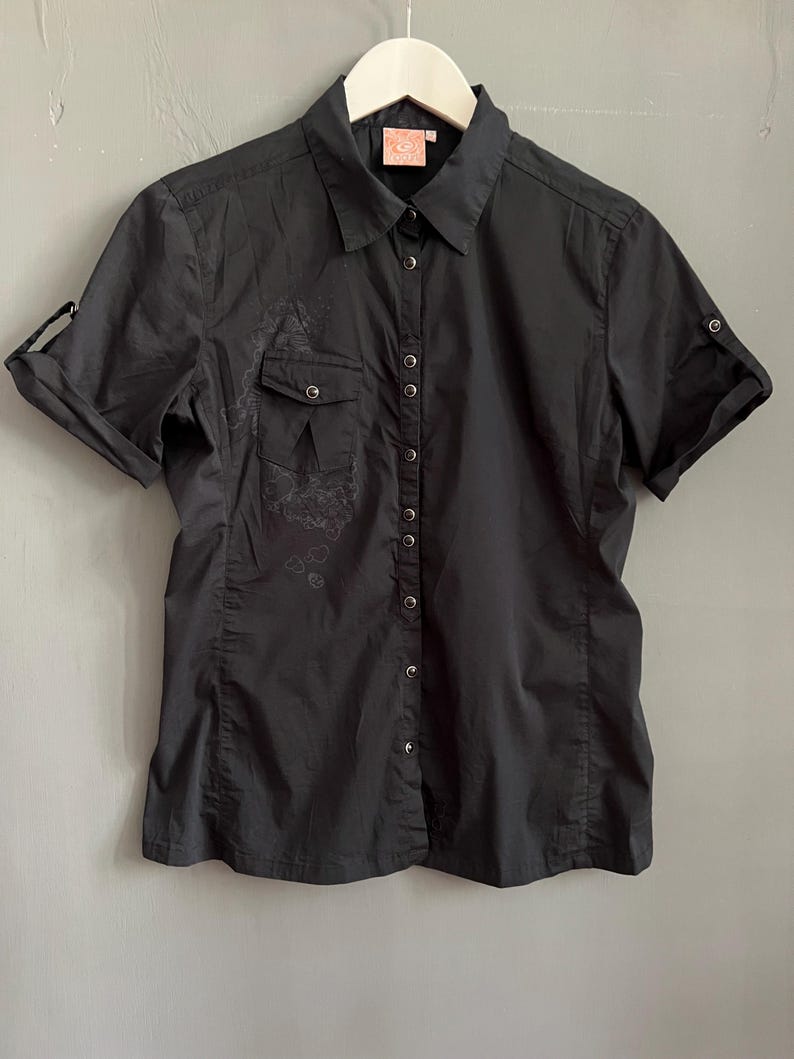 May include: Black short-sleeve button-down shirt with a collared neckline and a small pocket on the left chest. The shirt has a subtle floral design on the front and roll-up sleeves. The shirt is hanging on a white hanger.