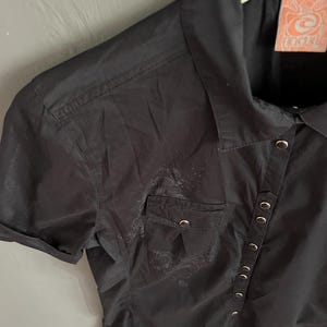 May include: Black short-sleeved button-up shirt with a small pocket on the chest. The shirt has silver-colored buttons and a decorative design near the pocket. The tag on the shirt has an orange and white design.