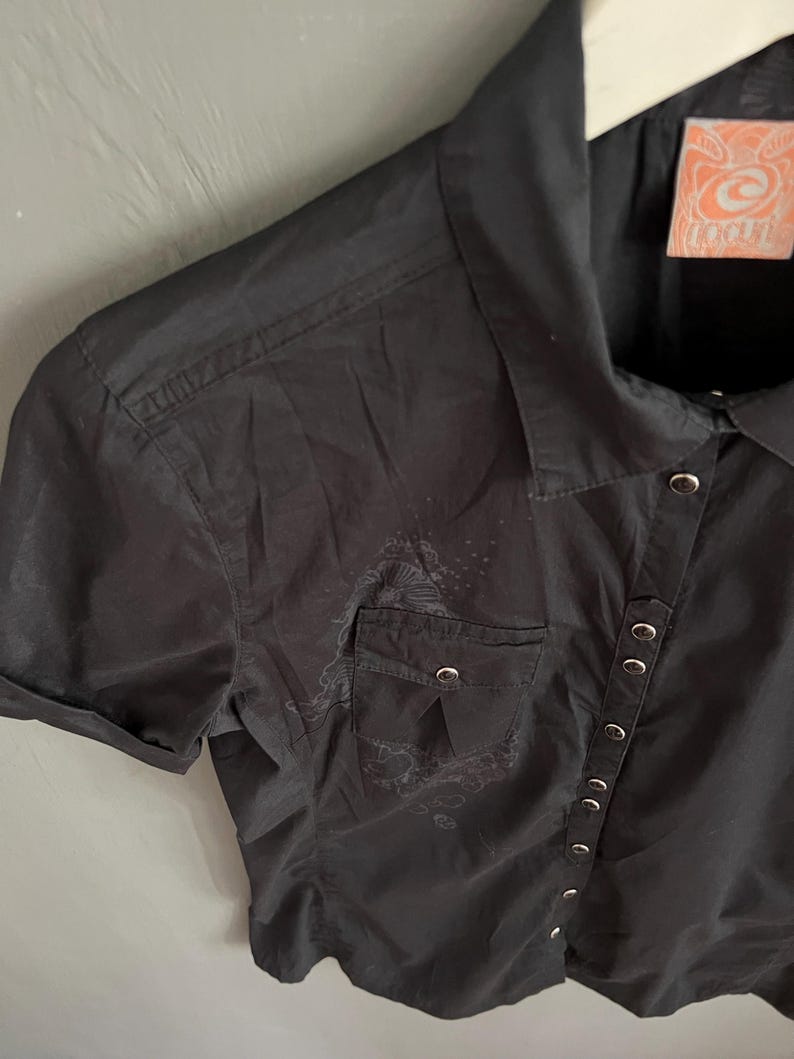 May include: Black short-sleeved button-up shirt with a small pocket on the chest. The shirt has silver-colored buttons and a decorative design near the pocket. The tag on the shirt has an orange and white design.