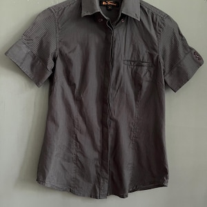May include: A dark grey short-sleeved shirt with vertical pinstripes, hanging on a white hanger. The shirt features a chest pocket and buttoned cuffs on the sleeves.