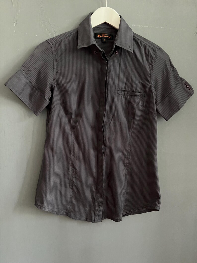 May include: A dark grey short-sleeved shirt with vertical pinstripes, hanging on a white hanger. The shirt features a chest pocket and buttoned cuffs on the sleeves.