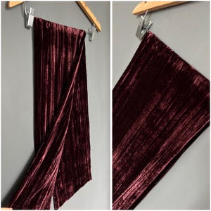 May include: A long, burgundy velvet scarf hangs from a wooden hanger against a grey wall. The scarf is folded, revealing its textured, pleated surface. The fabric has a rich, deep colour and a subtle sheen, suggesting a luxurious feel.